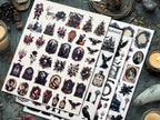 Two sheets of Halloween-themed fussy cut displayed on a dark, gothic tabletop with candles, a skull, and other spooky props. The designs include caskets, bats, haunted houses, bottles, keys, and portraits in a dark, vintage aesthetic.