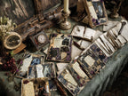 Flat lay of gothic-themed vintage stationery and journaling cards arranged on a dark tabletop with candles, keys, dried florals, and antique décor.