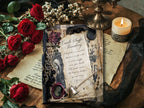 Moody product photo of a vintage-inspired gothic journal page with candle snuffer artwork, red roses, a lit pillar candle, black lace, wax seals, and layered antique parchment on a rustic wood surface.