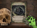 A styled flat-lay featuring a fantasy page titled 'Gods of the Necronomicon Tsathoggua' in the center, flanked by a realistic skull on the left and a green monster hand with black nails on the right, all set against a dark wood background.
