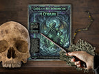 A tabletop scene featuring a printed page titled "Gods of the Necronomicon: Cthulhu" with fantasy artwork, placed on a dark wood surface. A human hand holds a wand near the card, and a skull plus dried mossy vine props are visible around it.