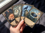 A hand holds four illustrated oracle or tarot-style cards against a dark fabric background near a window. The cards show fantasy scenes: a flower, a lantern in a bare tree, a raven on a rock, and a glowing well.