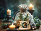 A silver drawstring gift bag filled with dried botanicals sits on a wooden surface, styled with lit candles, pinecones, and a decorative illustrated scroll.