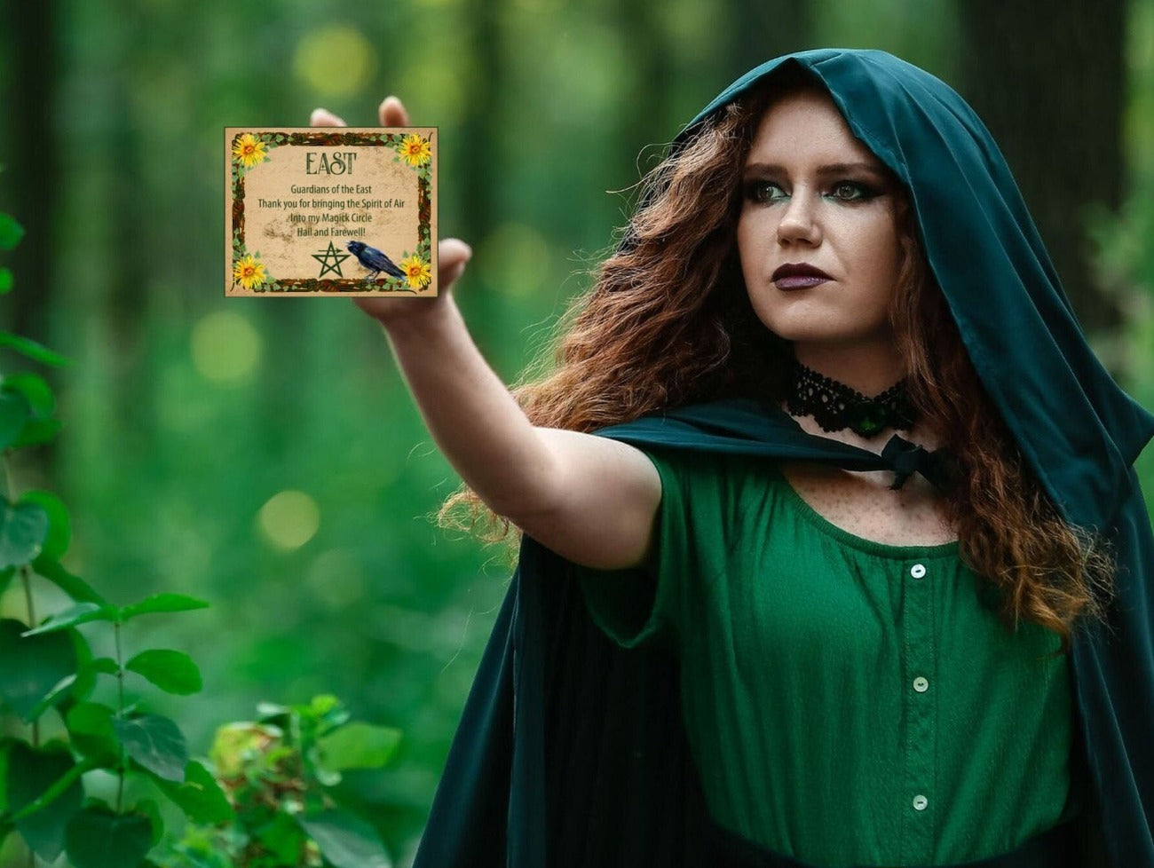 Wicca Woman dressed in a green cloak stands in a forest and holds out the East Invoking Quarter Card.